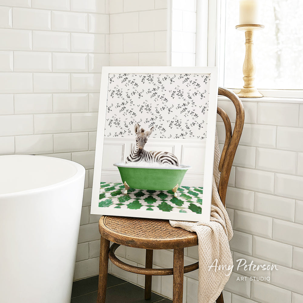 A framed image of a zebra sitting in a green bathtub is displayed on a wooden chair in a bathroom setting.