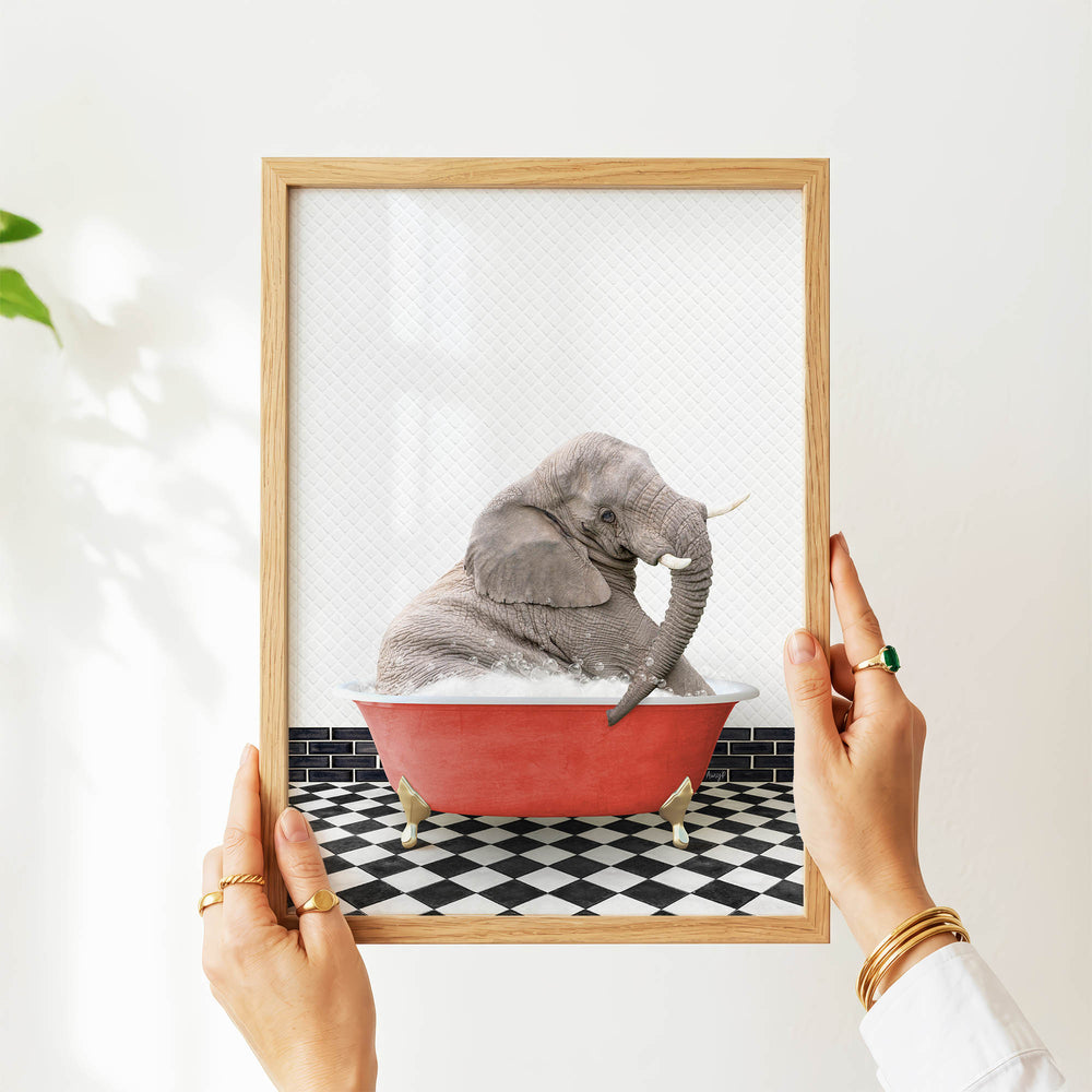 A person's hands are holding a framed picture of a gray elephant sitting in a red bathtub on a black and white checkered floor.