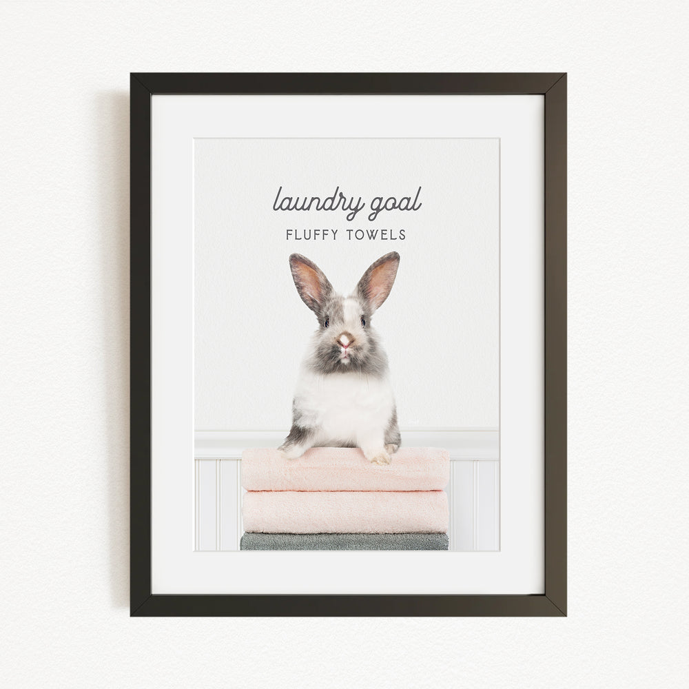 A framed image of a fluffy white rabbit sitting on a stack of pink towels, with the text "laundry goal fluffy towels" written above it.