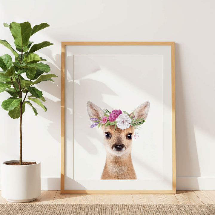 A framed print of a deer wearing a flower crown is displayed on a white wall, with a potted plant to the left.