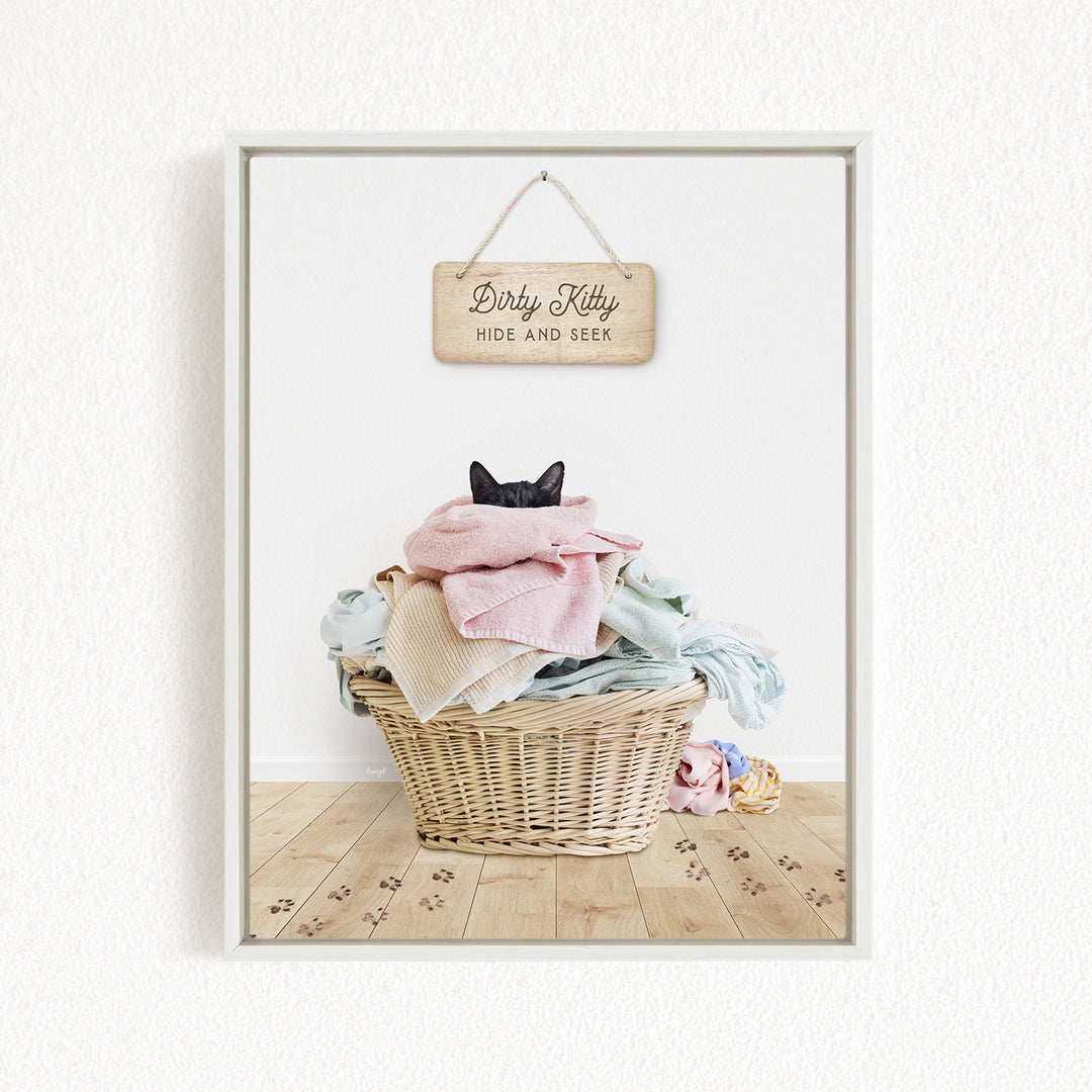 A black cat is sitting in a wicker basket filled with clothes, with a wooden sign above the basket that reads "Dirty Kitty HIDE AND SEEK".