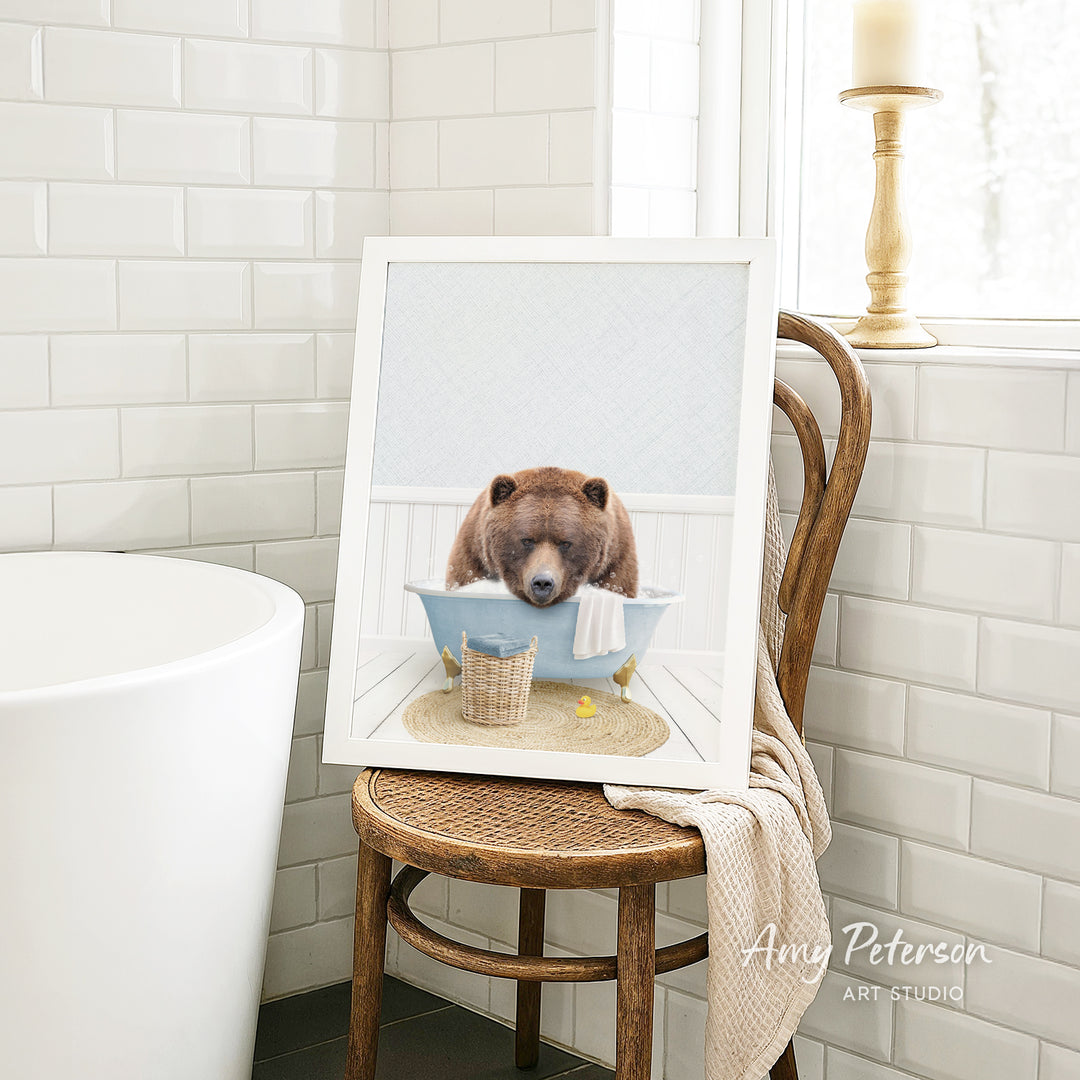 A framed image of a brown bear sitting in a bathtub, with a towel and basket nearby, is displayed on a wooden chair in a bathroom setting.