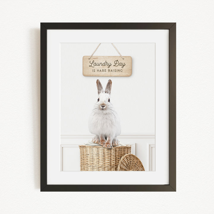 A framed image of a white rabbit sitting on a woven basket, with a wooden sign above it that says "Laundry Day is here raising".