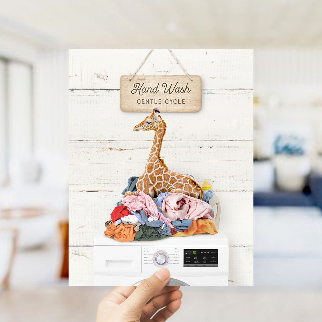 A hand holding a sign that says "Hand Wash Gentle Cycle" with a giraffe sitting on a pile of laundry underneath it.