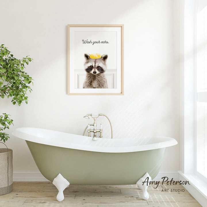 A framed print of a raccoon wearing a yellow crown is displayed on a white wall above a green clawfoot bathtub in a bright, airy bathroom.