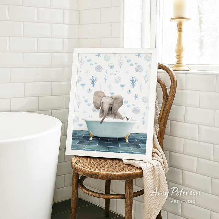 A framed image of an elephant sitting in a bathtub, surrounded by blue and white floral patterns, is displayed on a wooden chair in a bathroom setting.