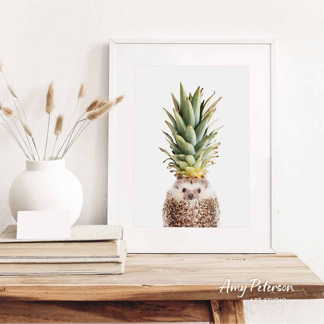 a framed print of a hedgehog in a pineapple, placed on a wooden shelf next to a white vase with dried flowers.