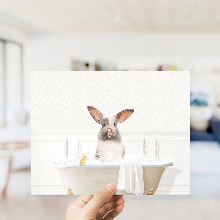 A hand holding a framed image of a rabbit sitting in a bathtub, with a small yellow bird perched on the edge of the tub.