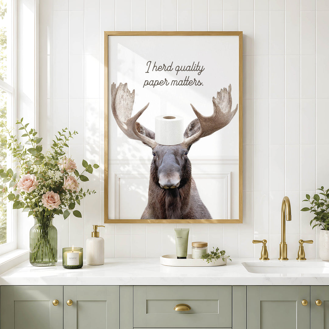 a framed picture of a moose with a roll of toilet paper on its head, placed above a bathroom sink with a vase of flowers and a candle on the counter.