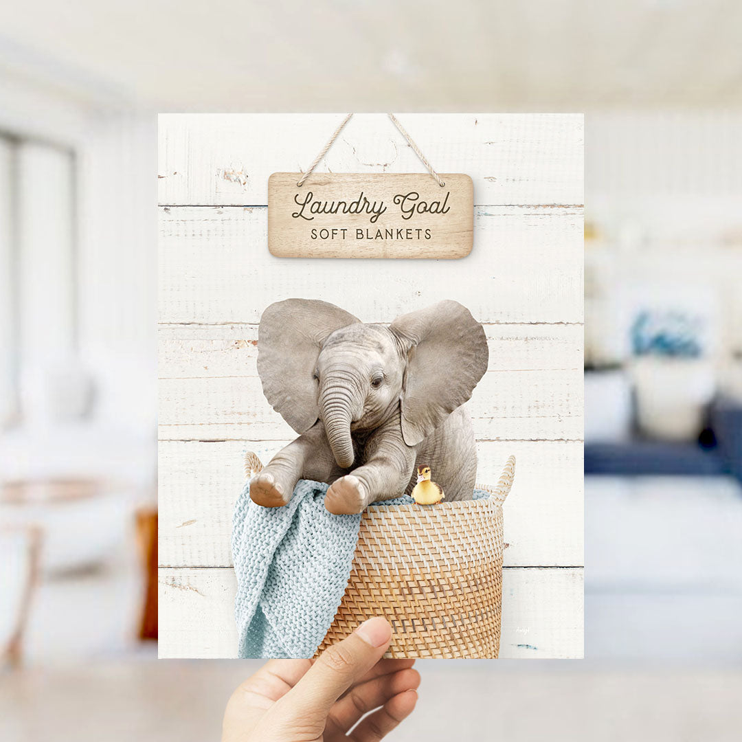 A hand is holding up a framed image of a baby elephant sitting in a woven basket, with a sign above it that says "Laundry Goal Soft Blankets".