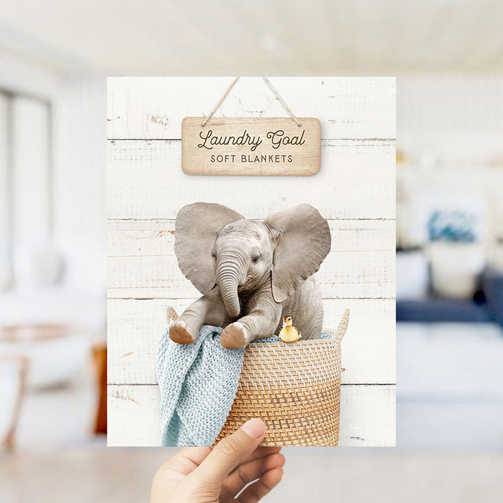 A hand is holding up a framed image of a baby elephant sitting in a woven basket, with a sign above it that says "Laundry Goal Soft Blankets".