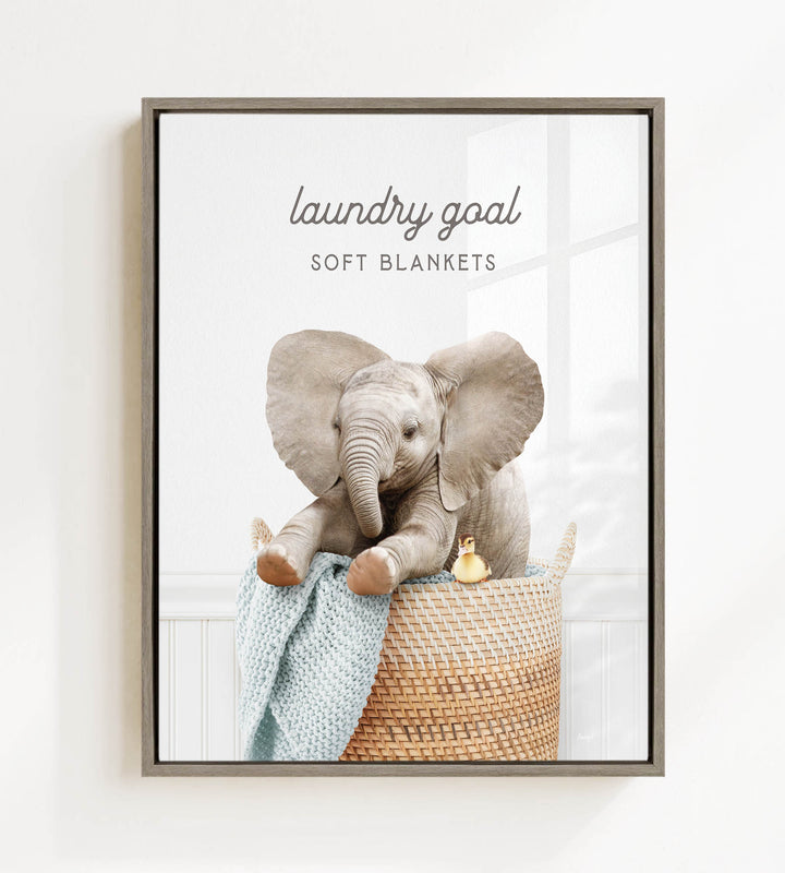 A framed image of a baby elephant sitting in a woven basket, with a blanket draped over its back.