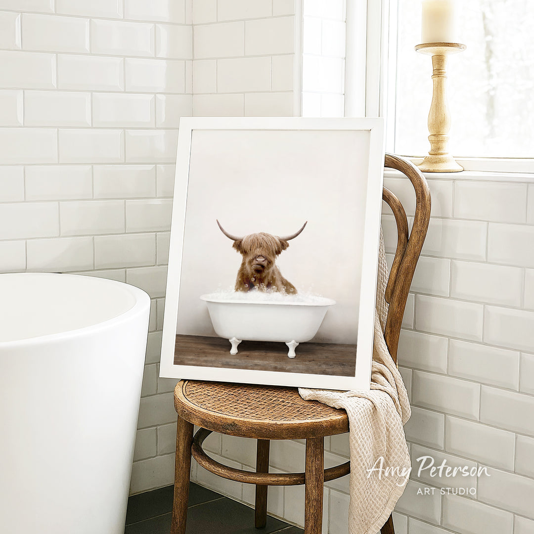 A framed image of a Highland cow sitting in a bathtub, with a towel draped over the back of a wooden chair in a bathroom setting.