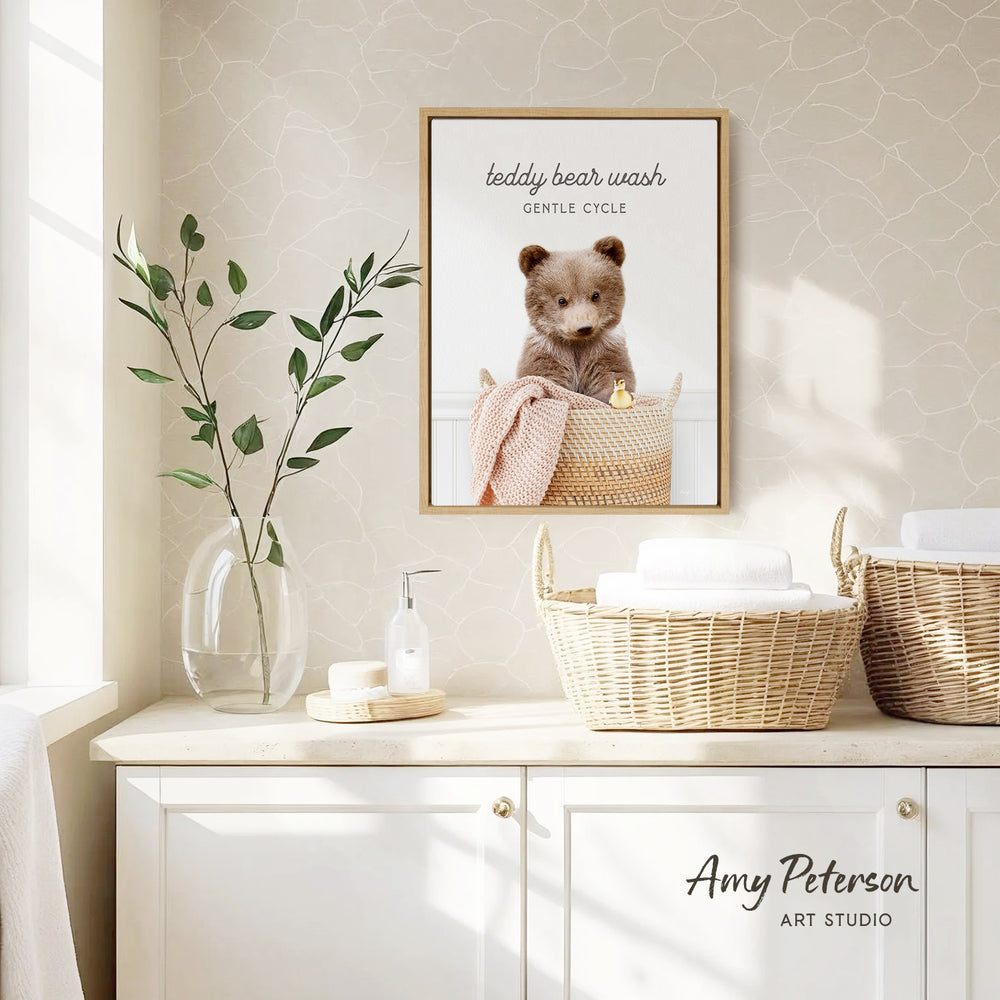 a framed photo of a teddy bear sitting in a basket, with the caption "teddy bear wash gentle cycle". The photo is displayed on a wall above a white cabinet, which has a vase of leaves and a bottle of soap on it.