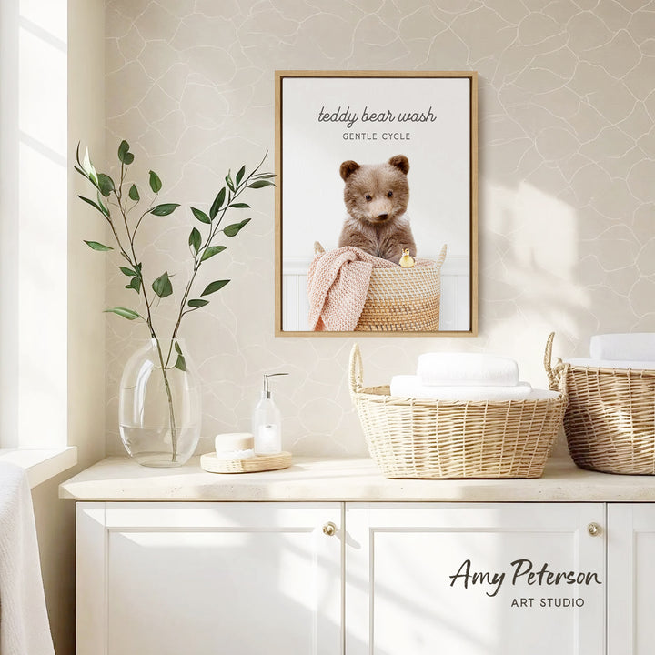 a framed photo of a teddy bear sitting in a basket, with the caption "teddy bear wash gentle cycle". The photo is displayed on a wall above a white cabinet, which has a vase of leaves and a bottle of soap on it.