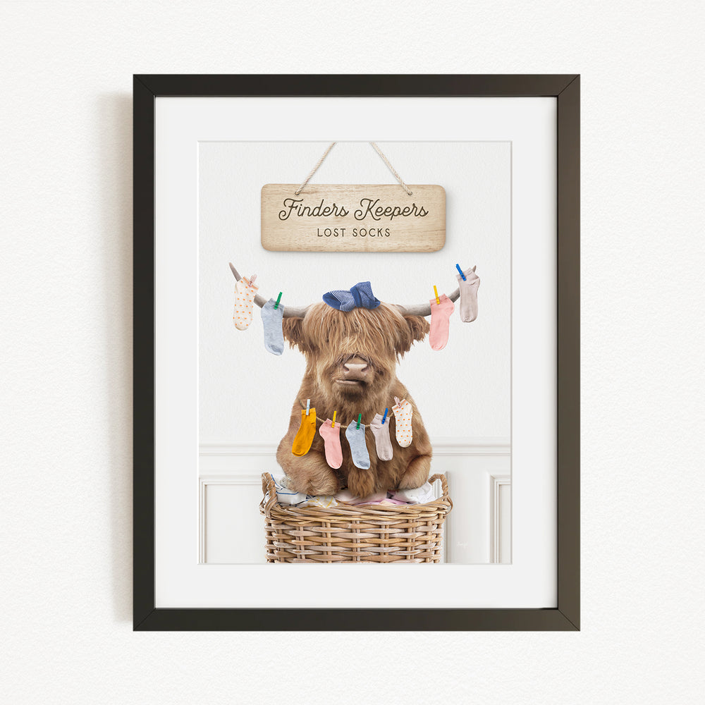 A framed image depicting a brown cow sitting in a wicker basket, with a sign above it that reads "Finders Keepers Lost Socks".