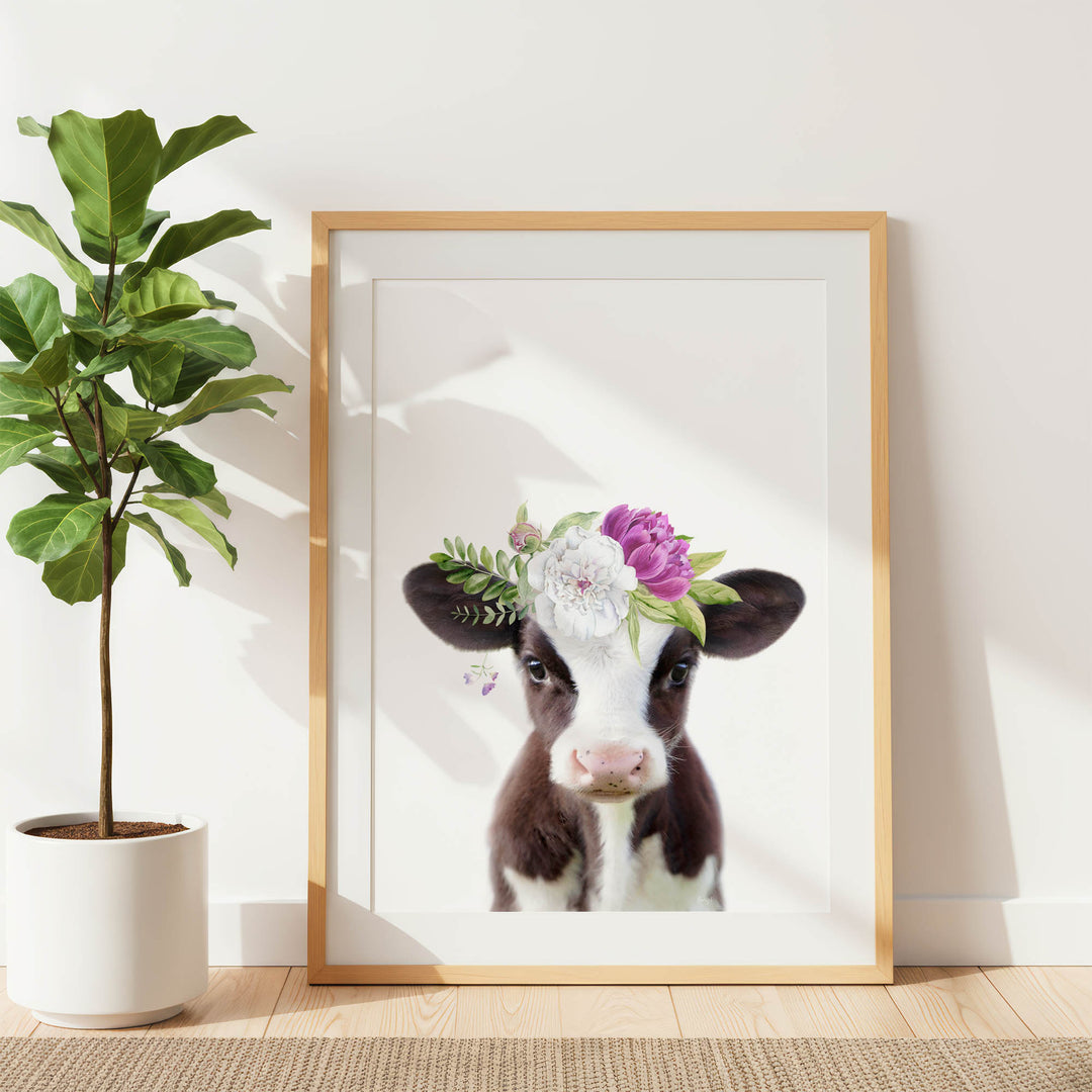 A framed print of a cow with a floral crown is displayed on a white wall, accompanied by a potted plant to the left.