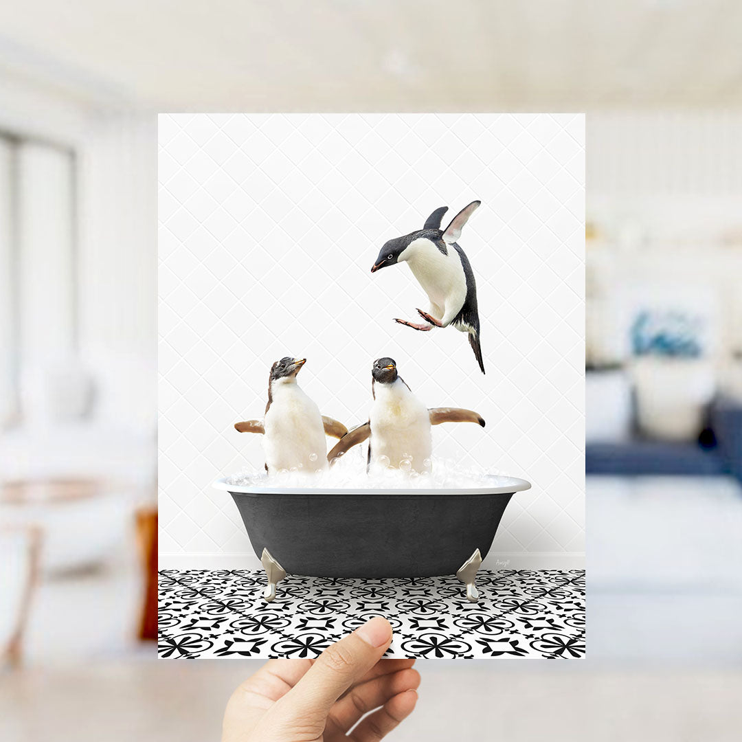 A hand holds up a framed photograph of two penguins sitting in a bathtub, with one penguin flying out of the tub.