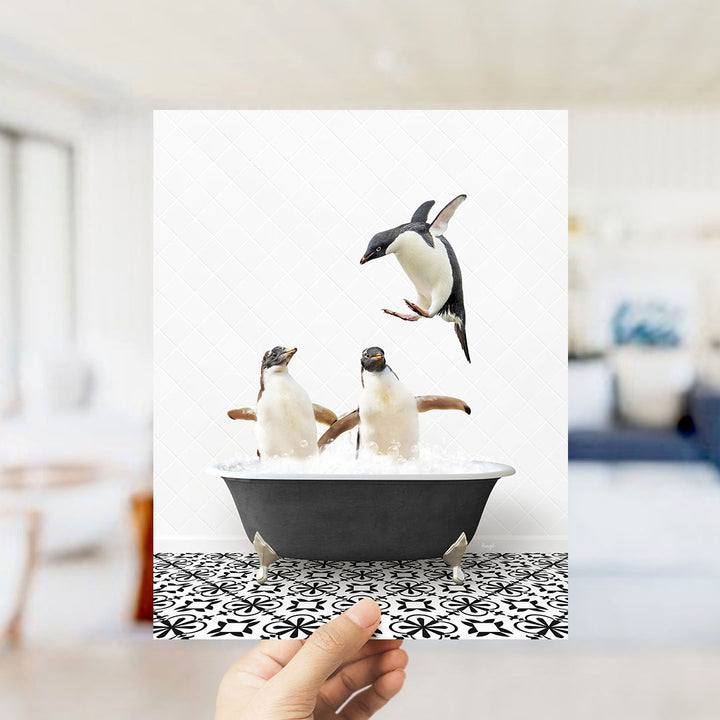 A hand holds up a framed photograph of two penguins sitting in a bathtub, with one penguin flying out of the tub.