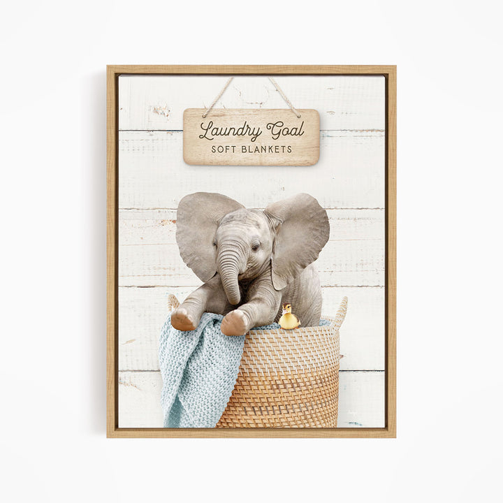 A framed image of a baby elephant sitting in a woven basket, with a sign above it that says "Laundry Goal Soft Blankets".