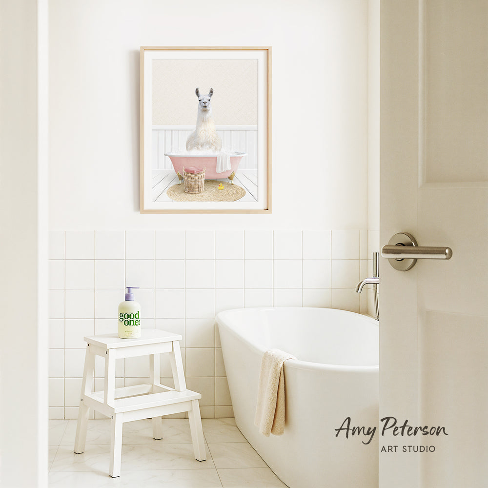 A framed image of a llama sitting in a pink bathtub is displayed on the wall above a white bathtub in a bathroom.