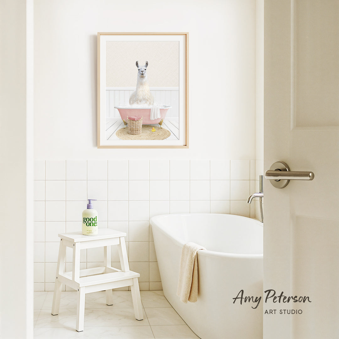 A framed image of a llama sitting in a pink bathtub is displayed on the wall above a white bathtub in a bathroom.