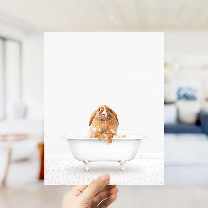 A hand holds up a framed image of a brown rabbit sitting in a white bathtub filled with water, creating a whimsical and playful scene.
