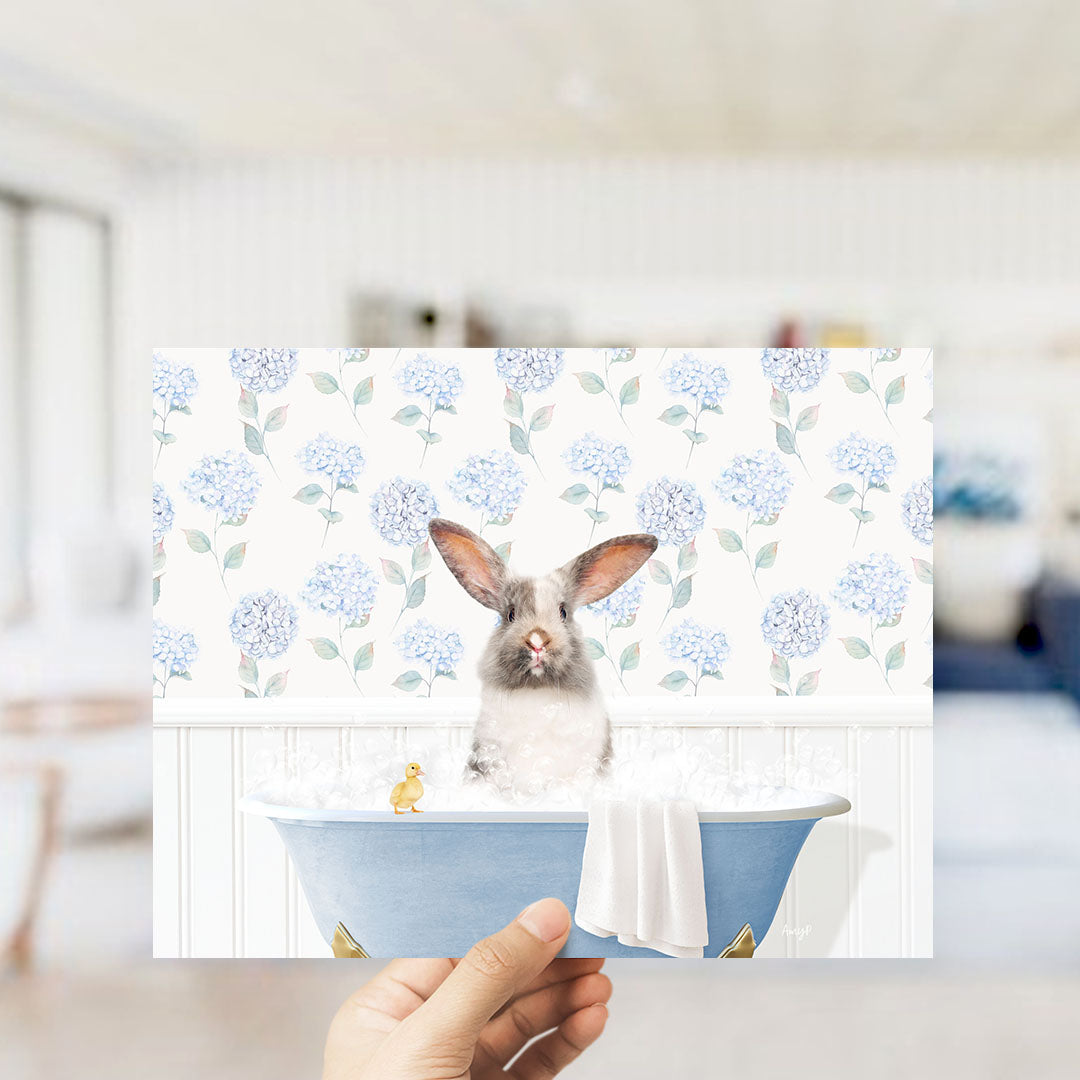 A hand holding a print of a rabbit sitting in a bathtub filled with water, surrounded by blue flowers.