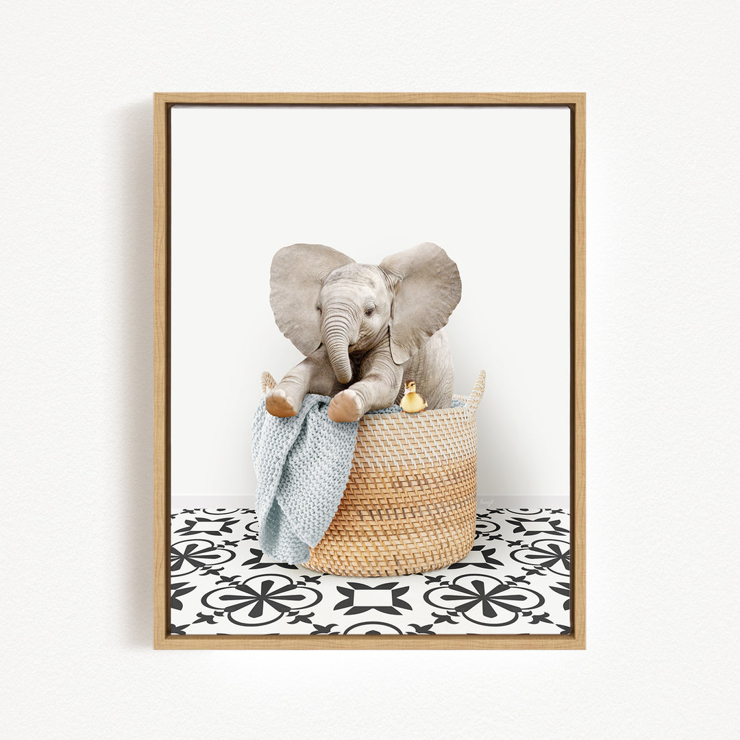 A gray elephant sits in a woven basket, holding a blue blanket and a yellow toy, against a white background with a black and white patterned floor.