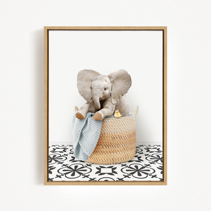 A gray elephant sits in a woven basket, holding a blue blanket and a yellow toy, against a white background with a black and white patterned floor.
