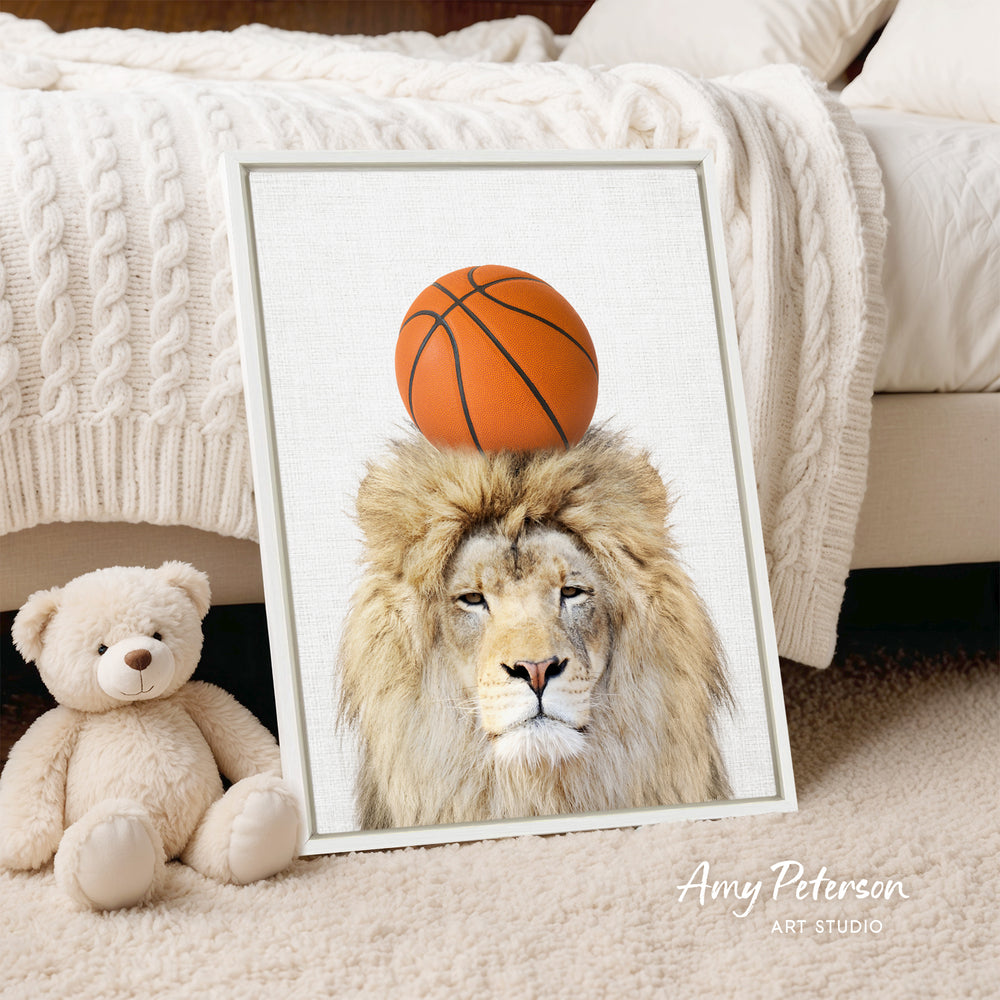 A framed image of a lion with a basketball on its head, placed on a bed next to a stuffed teddy bear.