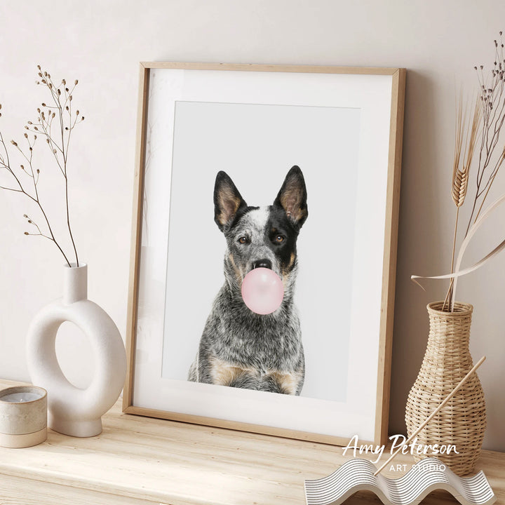 A framed photograph of a dog blowing a bubble gum bubble is displayed on a wooden shelf, accompanied by decorative vases and a woven basket.