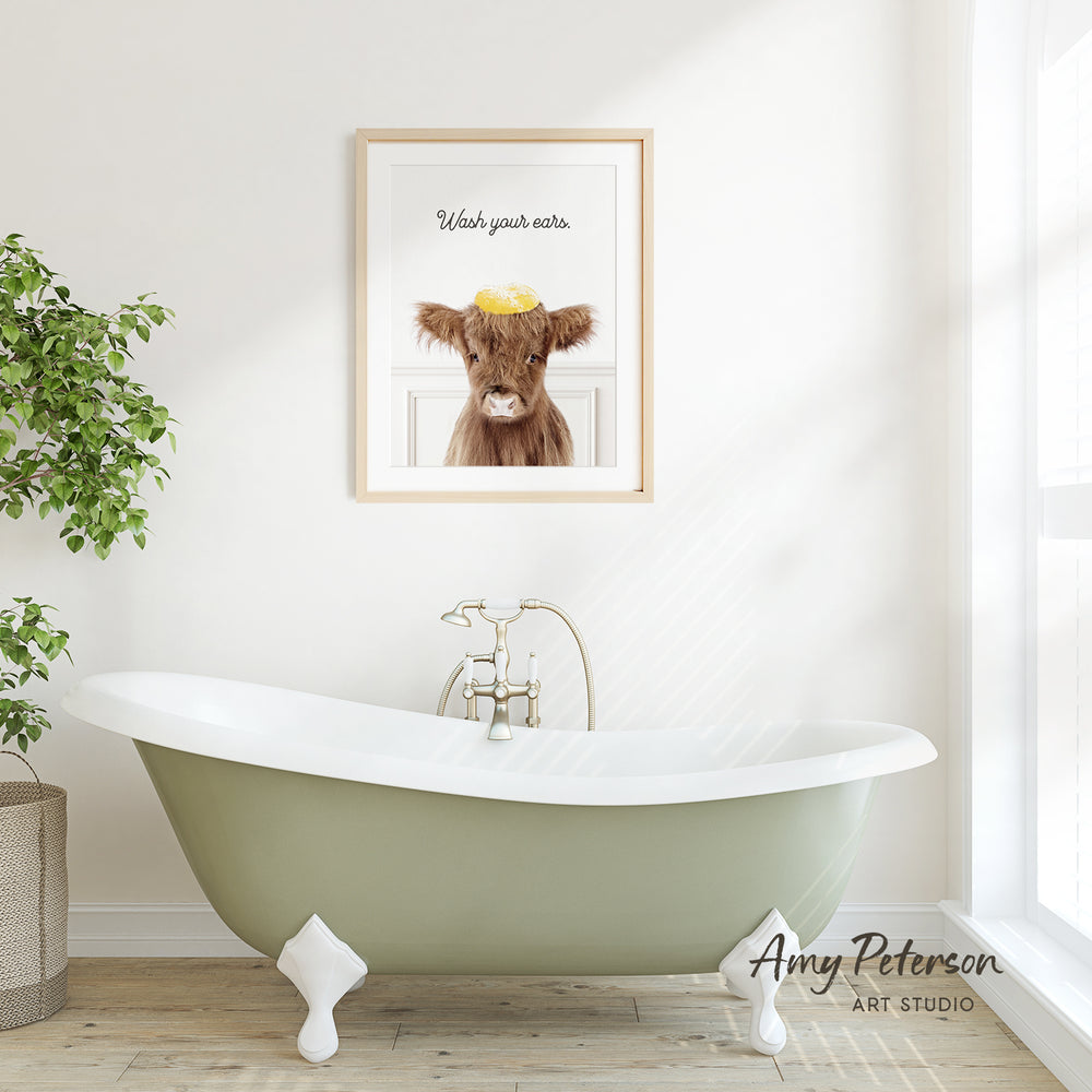 A framed image of a cow with a yellow hat is displayed on the wall above a green clawfoot bathtub in a bright, white bathroom.