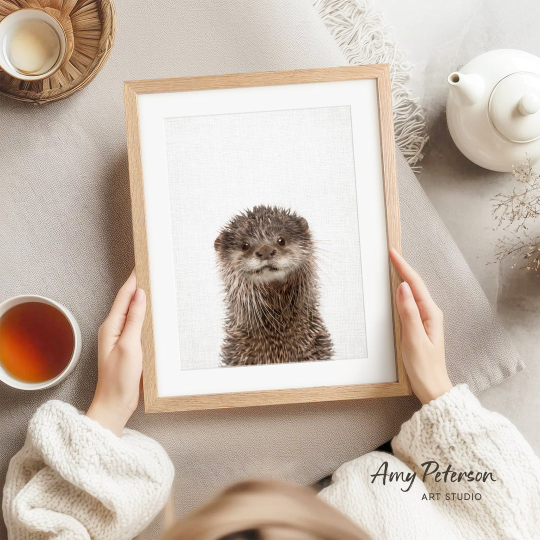 A person is holding a framed picture of an otter, which is a type of otter, in front of them. The person is wearing a white sweater and is sitting on a couch. There is a teapot, a cup, and a bowl on the couch, as well as a plant in the background.