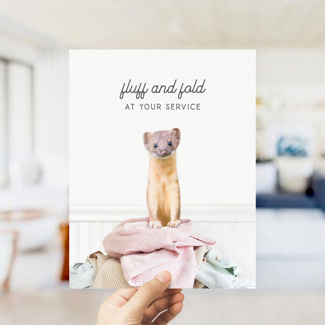 A hand holding a card with a photo of a small animal, likely a cat, sitting on a pile of clothes.