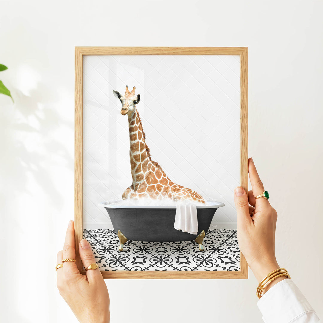 A person's hands are holding a framed picture of a giraffe sitting in a bathtub filled with water.