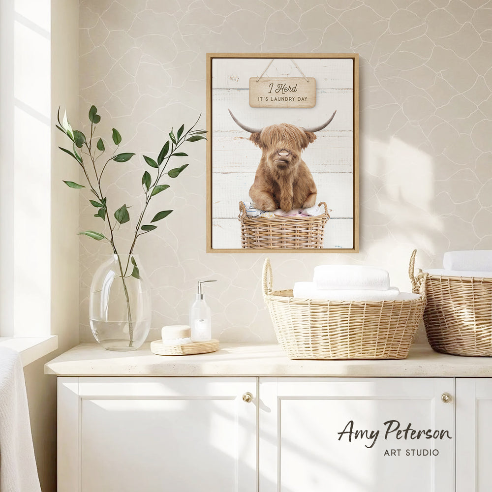 a framed picture of a Highland cow sitting in a wicker basket, with a sign above it that says "I herd it's laundry day". The picture is displayed on a wall above a white cabinet, which has a vase of greenery and a bottle of soap on it.