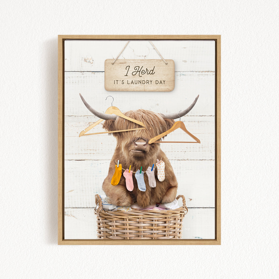 A framed image depicting a Highland cow sitting in a wicker basket, surrounded by clothes and a sign that reads "It's laundry day".