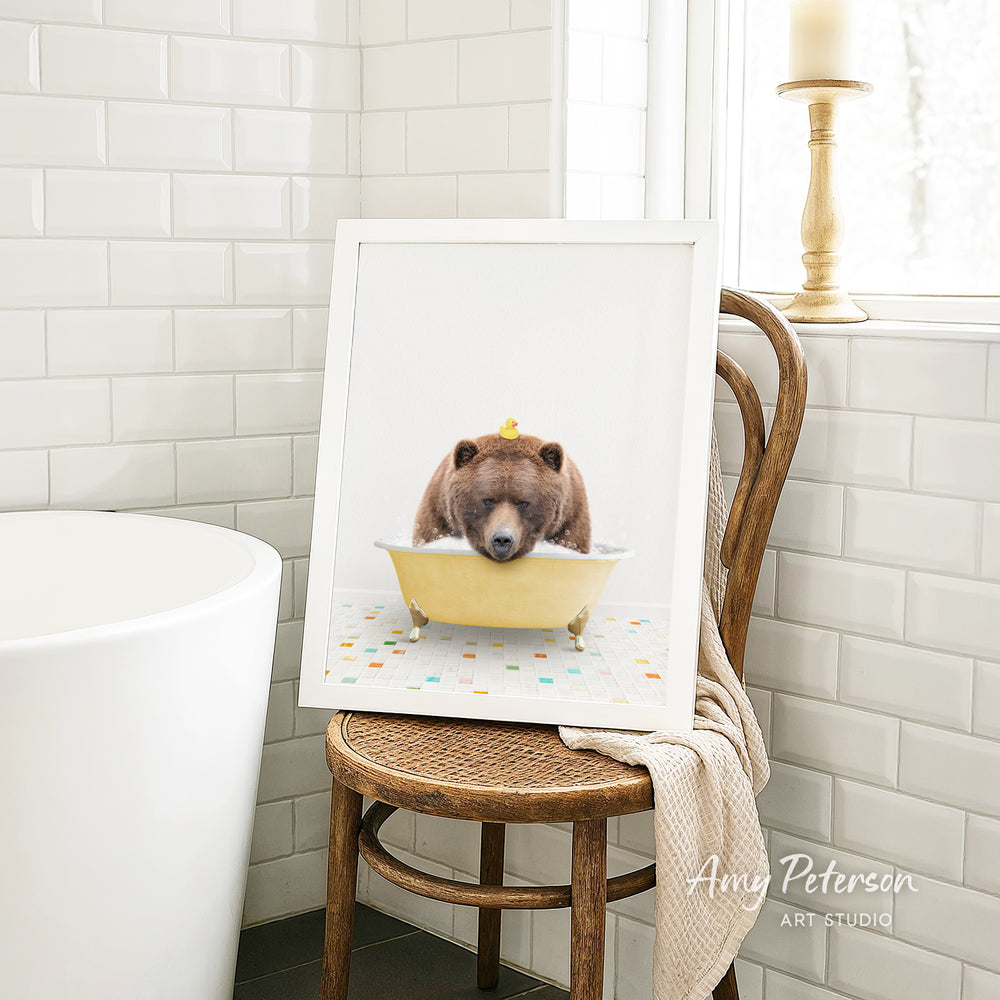 A framed image of a bear sitting in a bathtub, with a yellow bathtub and a towel draped over the back of a wooden chair.