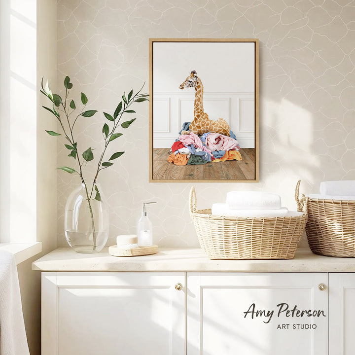 a giraffe sitting on a pile of clothes, with a framed print of the giraffe hanging on the wall above a wicker basket filled with towels.