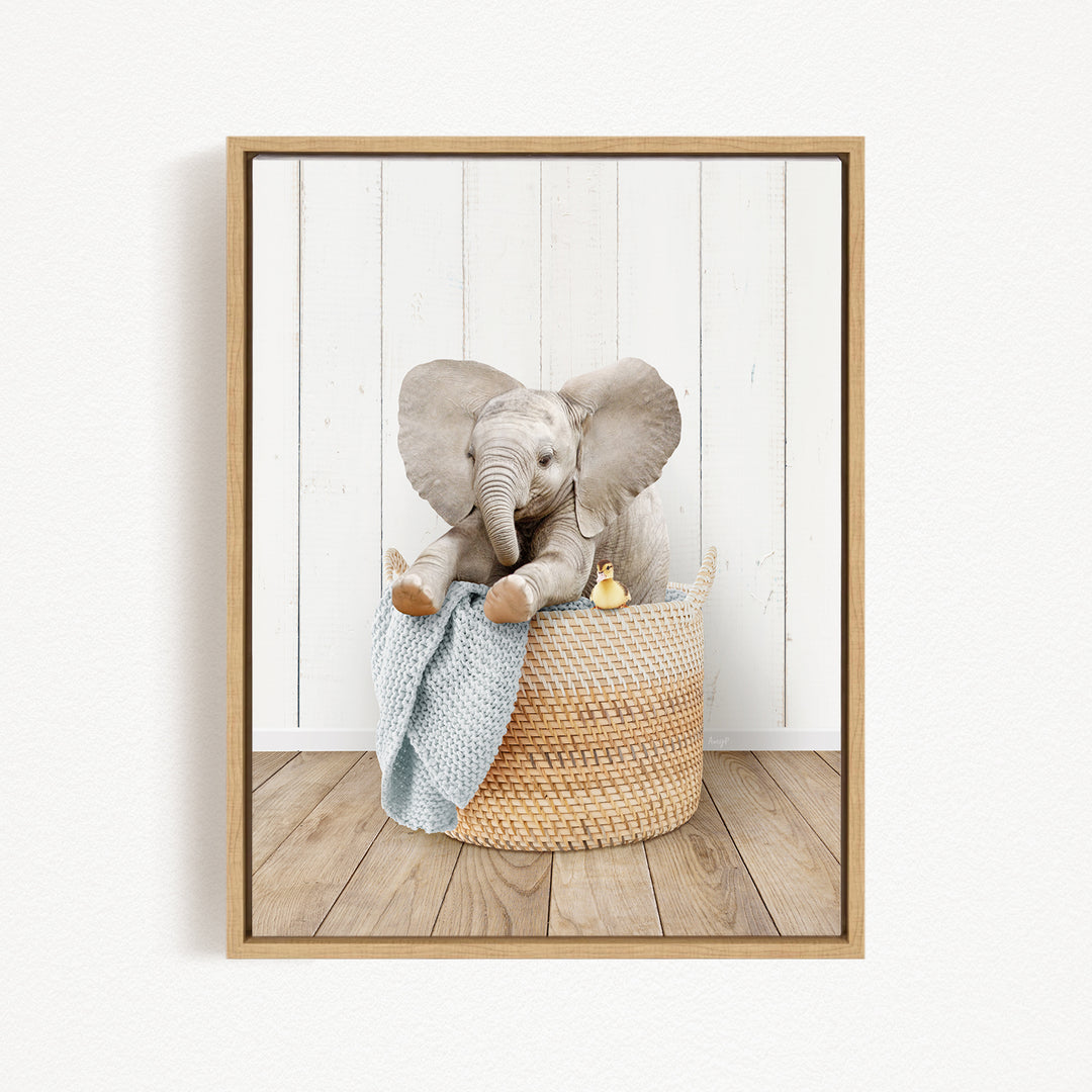 A gray elephant sits in a woven basket, holding a blue blanket and a yellow rubber duck, against a white wooden wall.