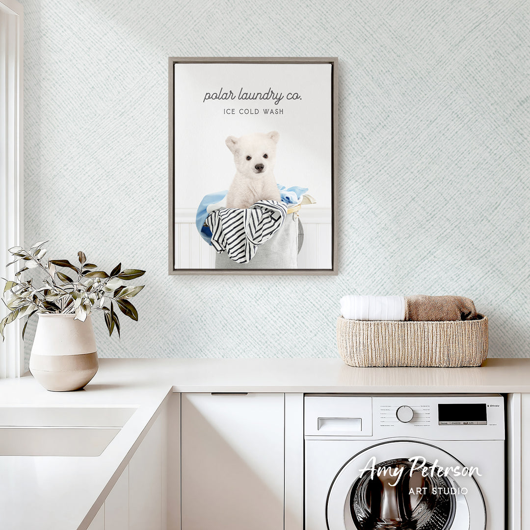 a framed poster of a polar bear sitting in a laundry basket, with the text "ice cold wash" and "polar laundry co." The poster is hanging on a wall above a white countertop with a plant and a basket of towels.
