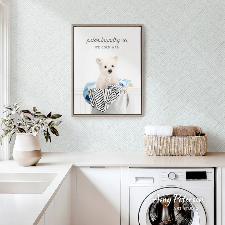 a framed poster of a polar bear sitting in a laundry basket, with the text "ice cold wash" and "polar laundry co." The poster is hanging on a wall above a white countertop with a plant and a basket of towels.