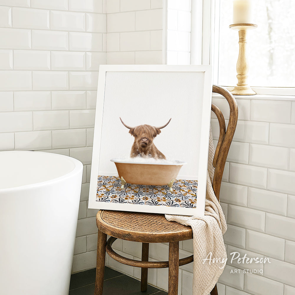 A framed image of a Highland cow sitting in a bathtub, with a towel draped over the back of a wooden chair in a bathroom setting.