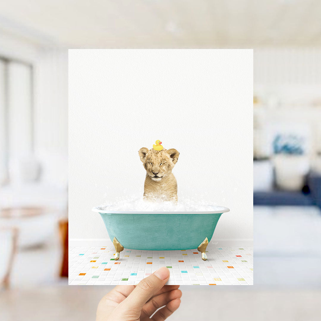 A hand holds up a framed image of a lion cub sitting in a bathtub filled with bubbles, with a yellow rubber duck perched on its head.