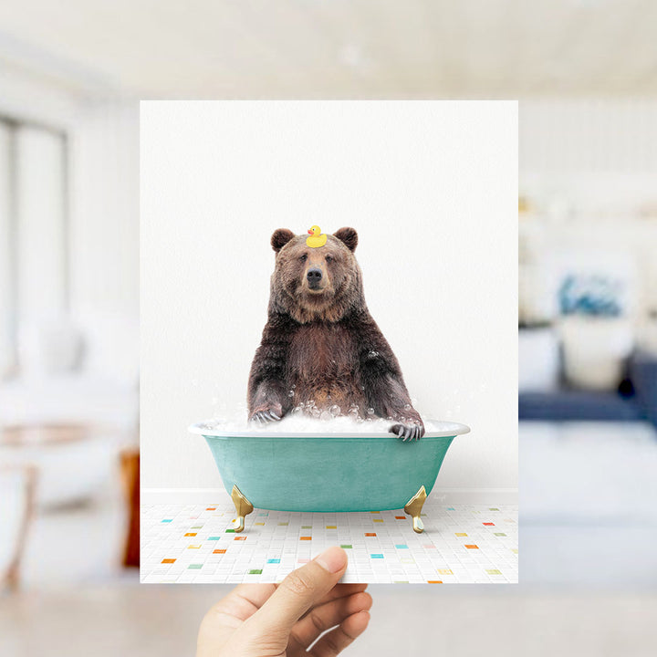 A hand holds up a framed image of a brown bear sitting in a green bathtub filled with bubbles, wearing a yellow rubber duck.