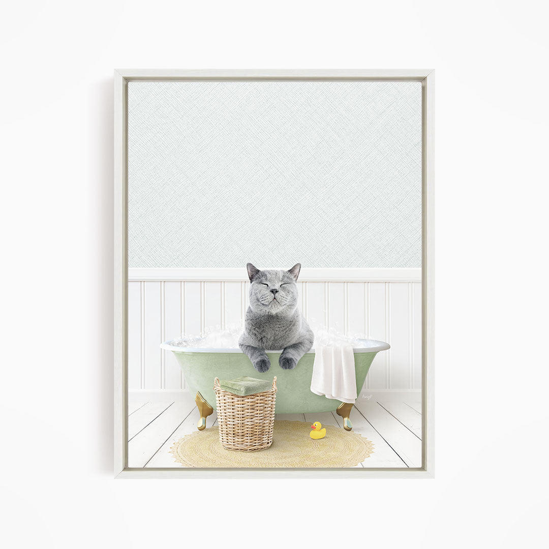 A gray cat sitting in a green bathtub filled with bubbles, with a yellow rubber duck nearby and a basket of bath toys on the floor.