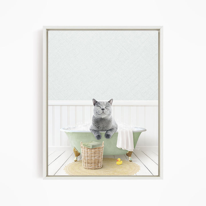 A gray cat sitting in a green bathtub filled with bubbles, with a yellow rubber duck nearby and a basket of bath toys on the floor.