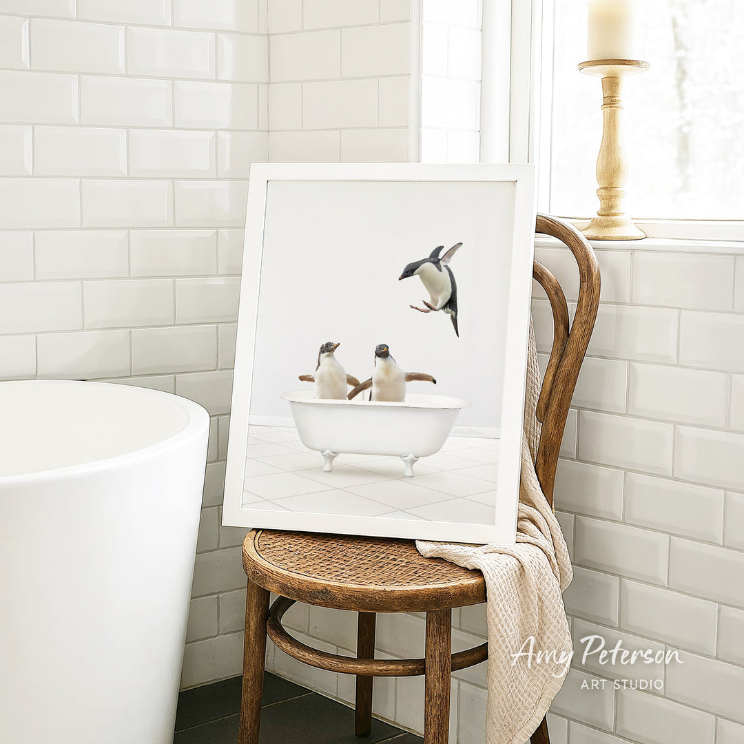 A framed image of three penguins sitting in a bathtub, with one penguin flying towards the others, is displayed on a wooden chair in a bathroom setting.
