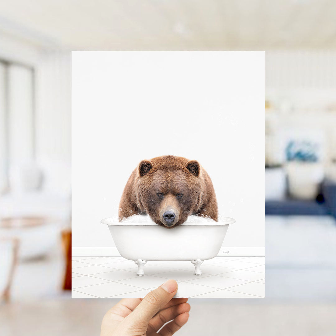 A hand holds up a framed image of a brown bear sitting in a bathtub filled with water.
